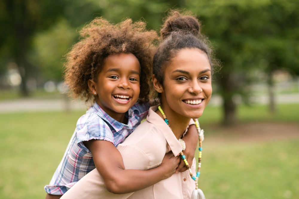 Young mom carrying child piggyback in park.