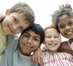 Group of four diverse children smiling and standing close together, looking down at the camera.