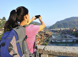 Female traveler with a backpack taking a photo of a scenic historic town and river from an elevated viewpoint.