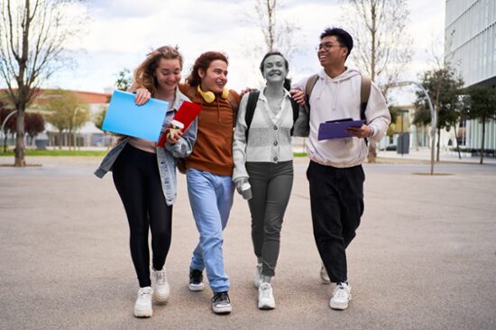 Four students walking on campus with arms around each other. One of them is greyed out. 